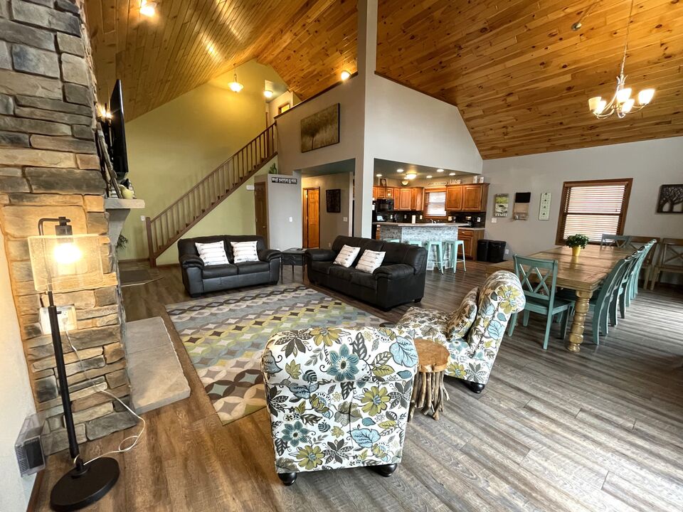 An open concept living space is showcased, featuring a cozy seating area with dark sofas and patterned chairs. A large dining table for 14 is positioned nearby, and décor includes natural wood accents, high ceilings, and a stone fireplace, providing a warm and welcoming environment.