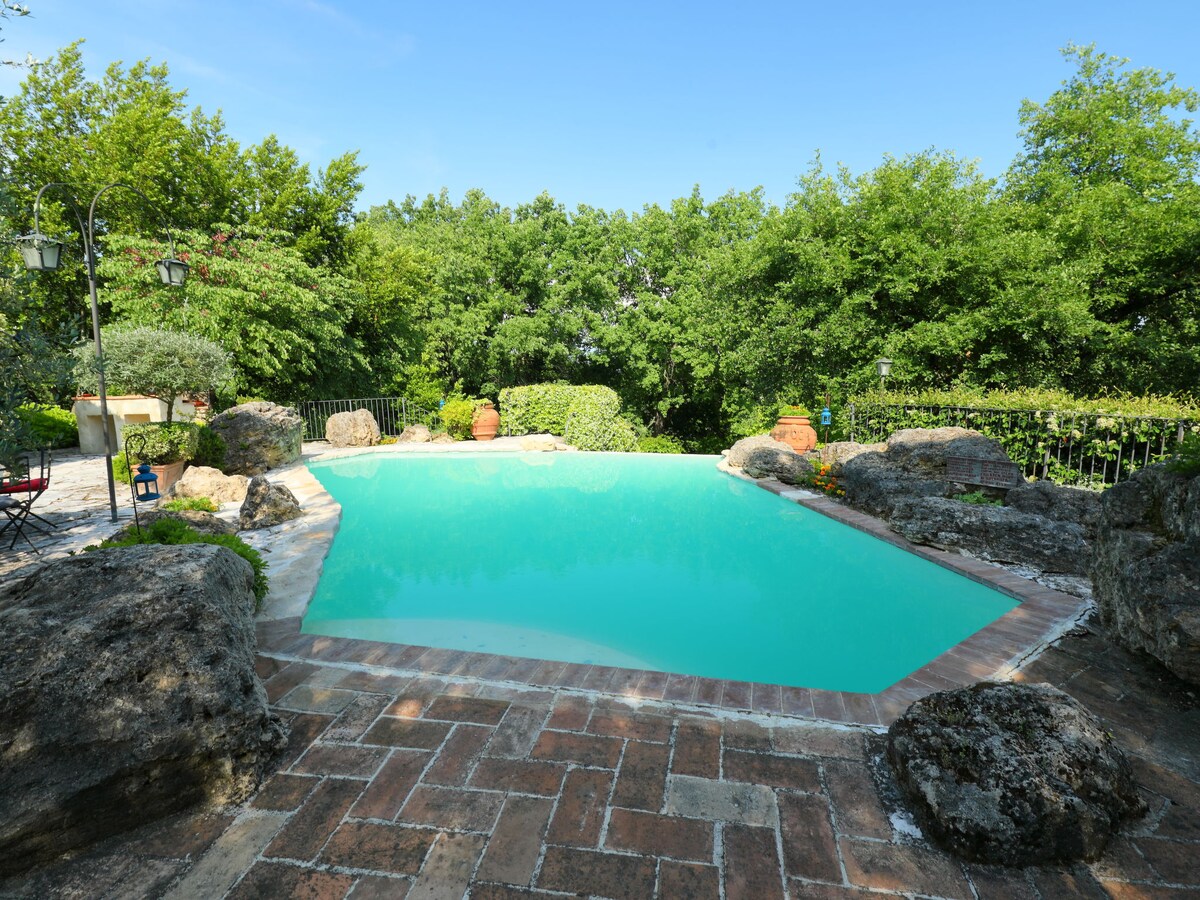 A uniquely shaped swimming pool is surrounded by natural stone and a patio area. Lush greenery provides privacy and enhances the serene setting. Sunlight reflects off the clear water, inviting relaxation amidst the tranquil outdoor space.