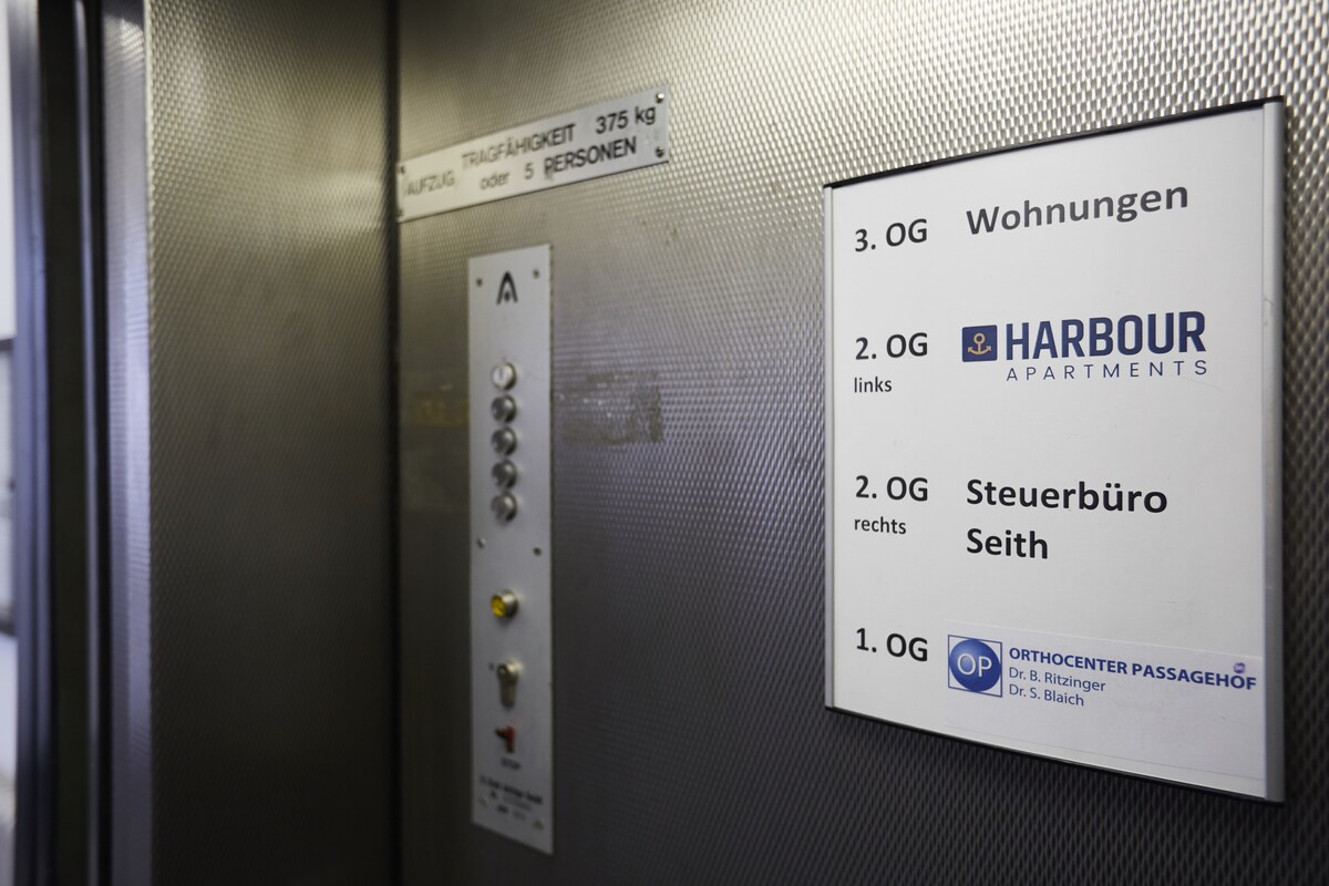 An elevator panel is shown, featuring a metallic finish. Control buttons are visible, alongside a list of floors including 'HARBOUR APARTMENTS' on the second floor. Clear directional signs indicate nearby offices and amenities, contributing to a functional and accessible environment.