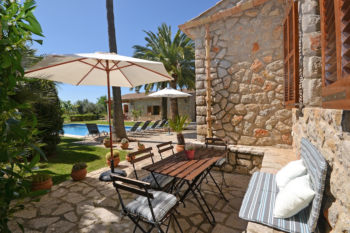A stone terrace features a wooden dining table surrounded by four folding chairs, with cushions for comfort. An umbrella provides shade, while palm trees and a carefully maintained garden frame the private pool in the background, enhancing the outdoor dining experience.