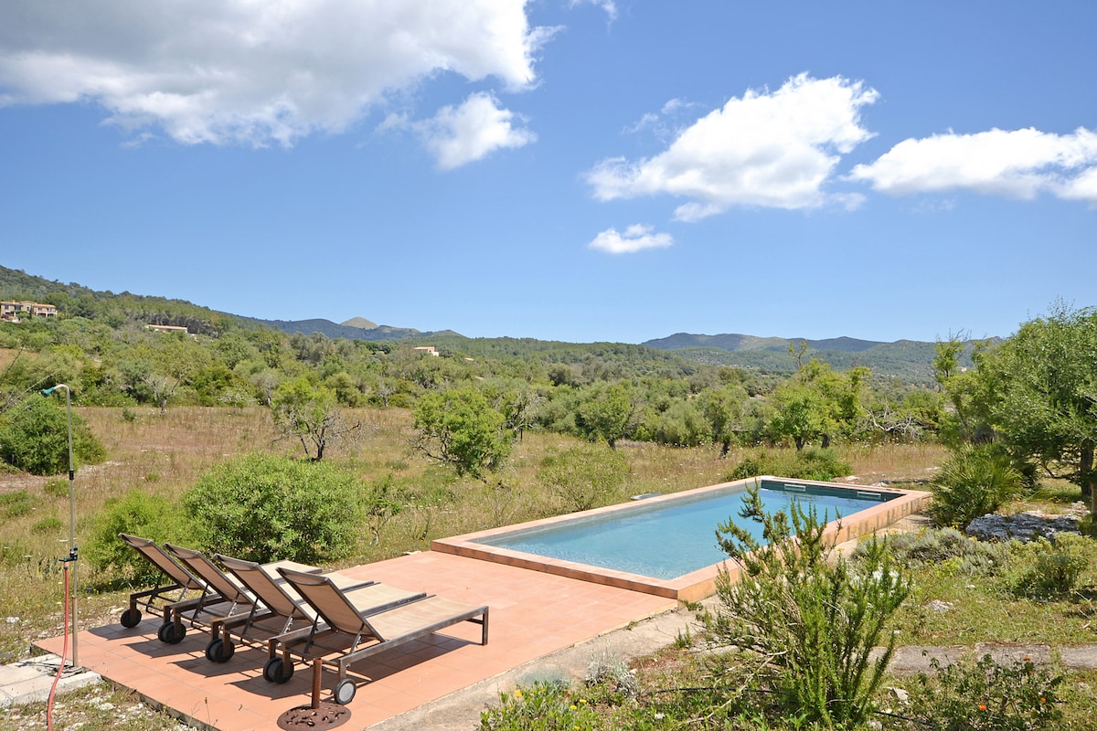 A serene outdoor area features a rectangular swimming pool bordered by a sun terrace. Four lounge chairs are arranged along one side, providing views of the surrounding greenery and distant hills under a bright blue sky with scattered clouds.
