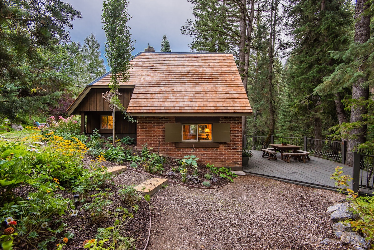 A charming cottage is framed by lush greenery, featuring a sloped roof and brick exterior. Colorful flowers bloom in the foreground, while a wooden deck with a picnic table provides outdoor seating. Large windows invite natural light into the cozy interior.