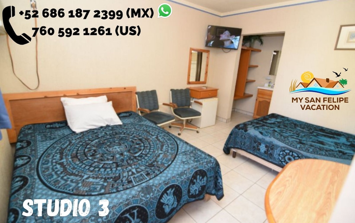 A studio space features two beds covered with patterned blue textiles. A mirror and dressing area are visible, along with a shelf and seating. A small television is mounted on the wall, and light flows in through a window, illuminating the tiled floor.
