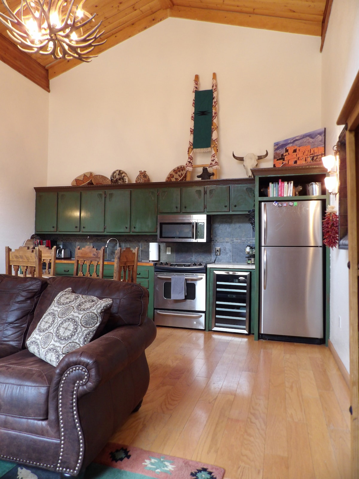A modern kitchen area is equipped with green cabinets and stainless steel appliances, including a refrigerator and a stove. The living space features a brown leather sofa adorned with decorative pillows, while warm wood accents and high ceilings create an open and inviting atmosphere.