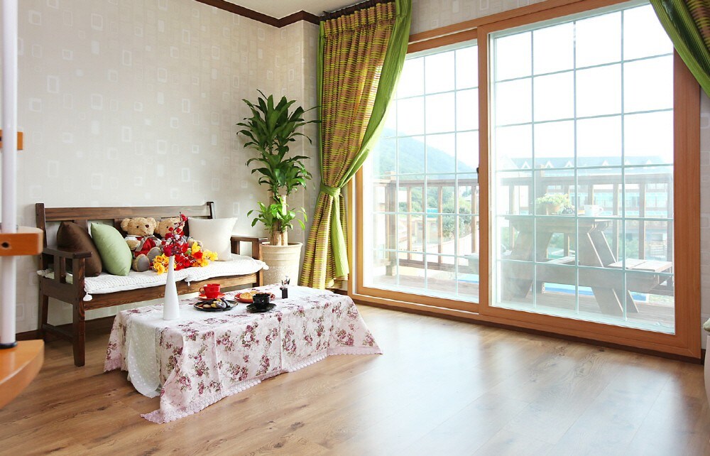 A bright living area is featured with large windows that provide views of the surrounding landscape. A wooden bench adorned with plush toys and colorful cushions sits beside a low table set with decorative items. Natural light floods the space, complemented by green curtains.