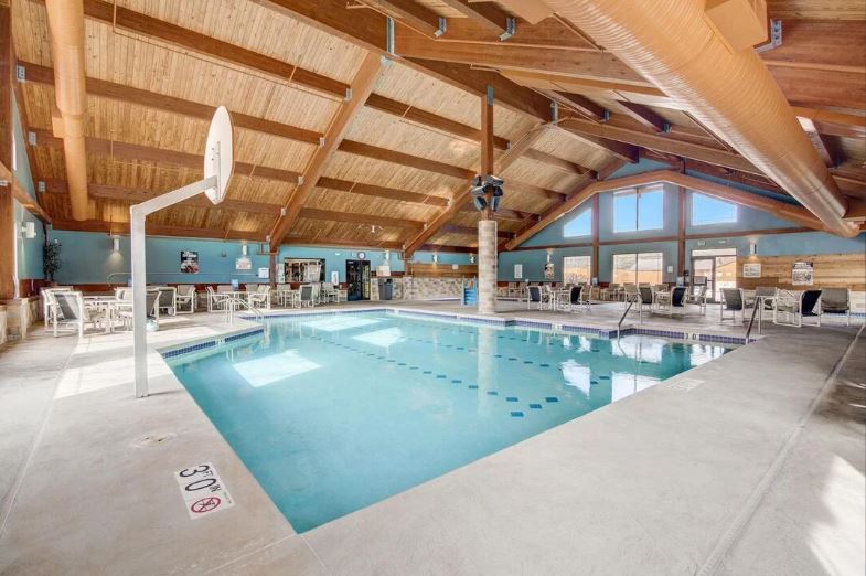 An indoor pool area features a clear blue pool under a vaulted wooden ceiling. Sunlight streams through large windows, enhancing the spacious environment. Surrounding the pool are multiple lounge chairs and a basketball hoop, creating a versatile space for relaxation and recreation.