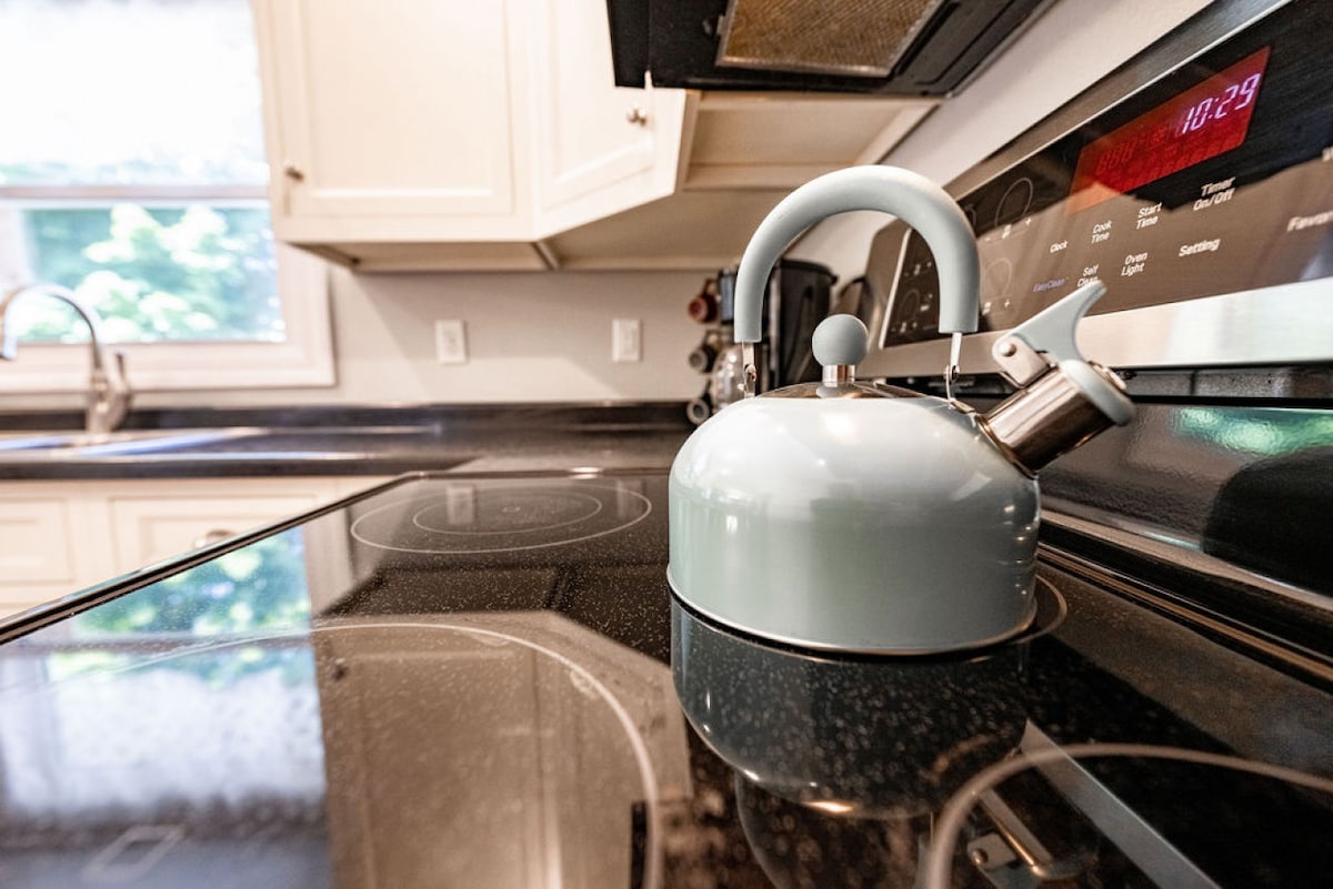 A pastel blue kettle is placed on a modern black stovetop, reflecting the surrounding kitchen's light. The cabinetry features light-colored doors, and a window is partially visible, allowing natural light to enter the space.