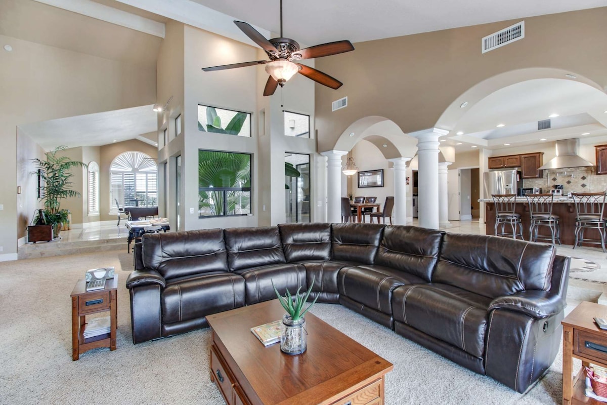 A spacious living area features a large sectional couch, coffee table, and tasteful decor. High ceilings with a ceiling fan and large windows allow for ample natural light, while columns create an open and inviting flow to the adjoining kitchen and dining area.