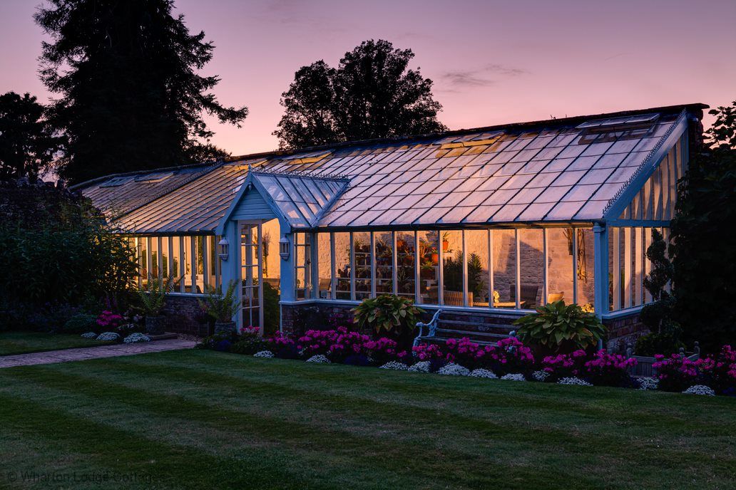 A spacious glasshouse is showcased at dusk, illuminated from within. The structure features large glass panels and a peaked roof, surrounded by well-maintained gardens and vibrant flower beds. Lush greenery complements the serene setting, enhancing the peaceful atmosphere of the countryside.