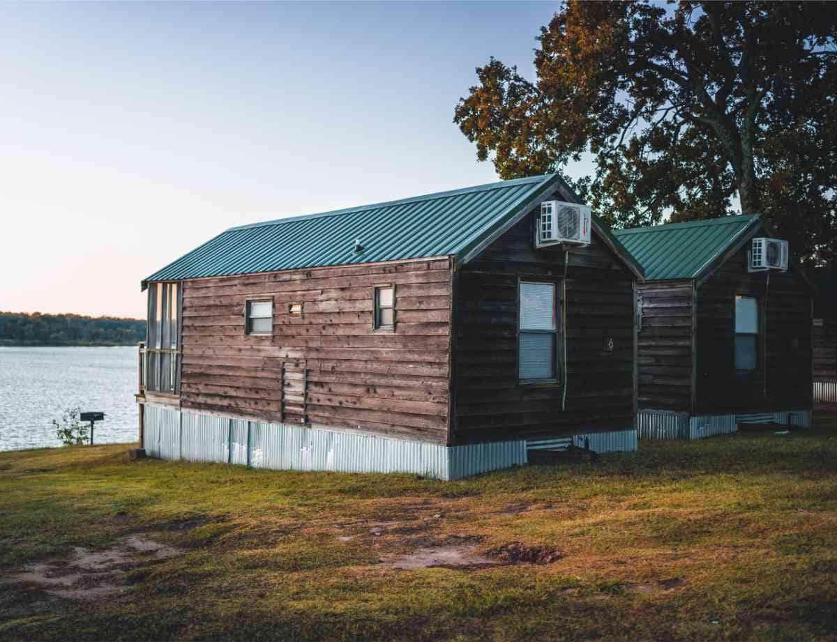 Two rustic cabins with wooden exteriors are positioned beside a serene lake. Each cabin features a metal roof and air conditioning units. The grassy area around the cabins provides a natural setting, enhancing the peaceful lakeside environment.