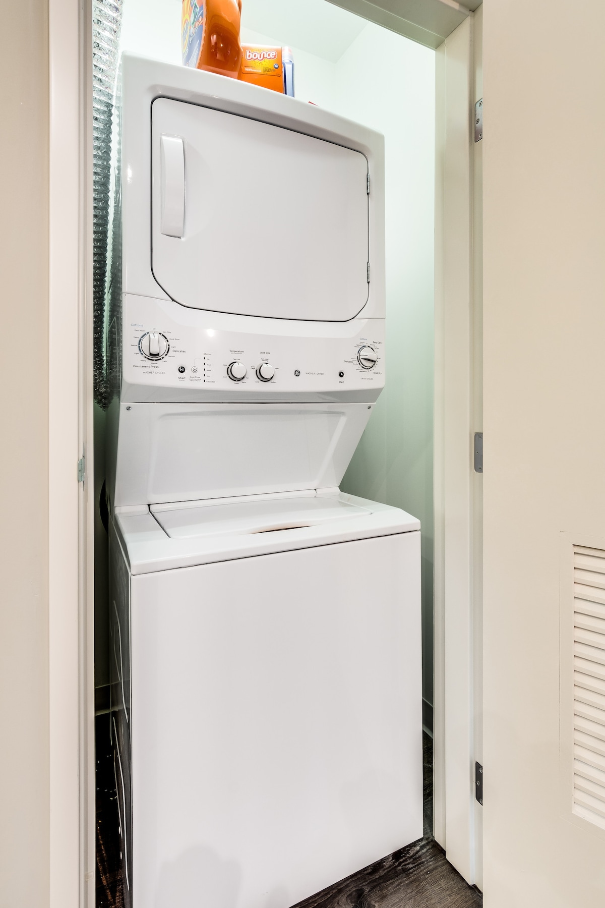 A compact laundry area features a stacked washer and dryer in white. Shelves above hold a container of laundry detergent. The space is enclosed by a door, ensuring a neat and organized appearance.