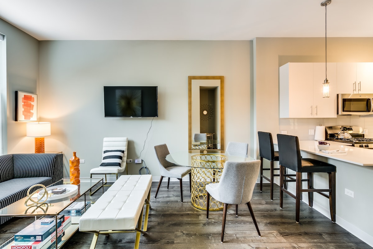 A modern living space features a combination of comfortable seating arrangements, including a sleek sofa and various chairs. Bright natural light floods the area, highlighting a stylish dining table and a mounted television. Contemporary art and decorative elements add to the inviting ambiance.