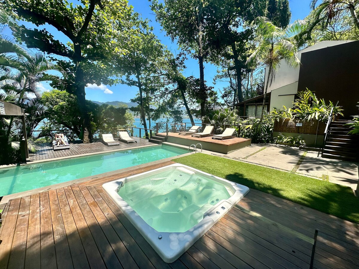 The outdoor area features a wooden deck with a hot tub and a swimming pool, surrounded by tropical greenery. Sun loungers are positioned nearby, offering views of the water. The setting is bright, with clear blue skies and gentle sunlight filtering through the trees.
