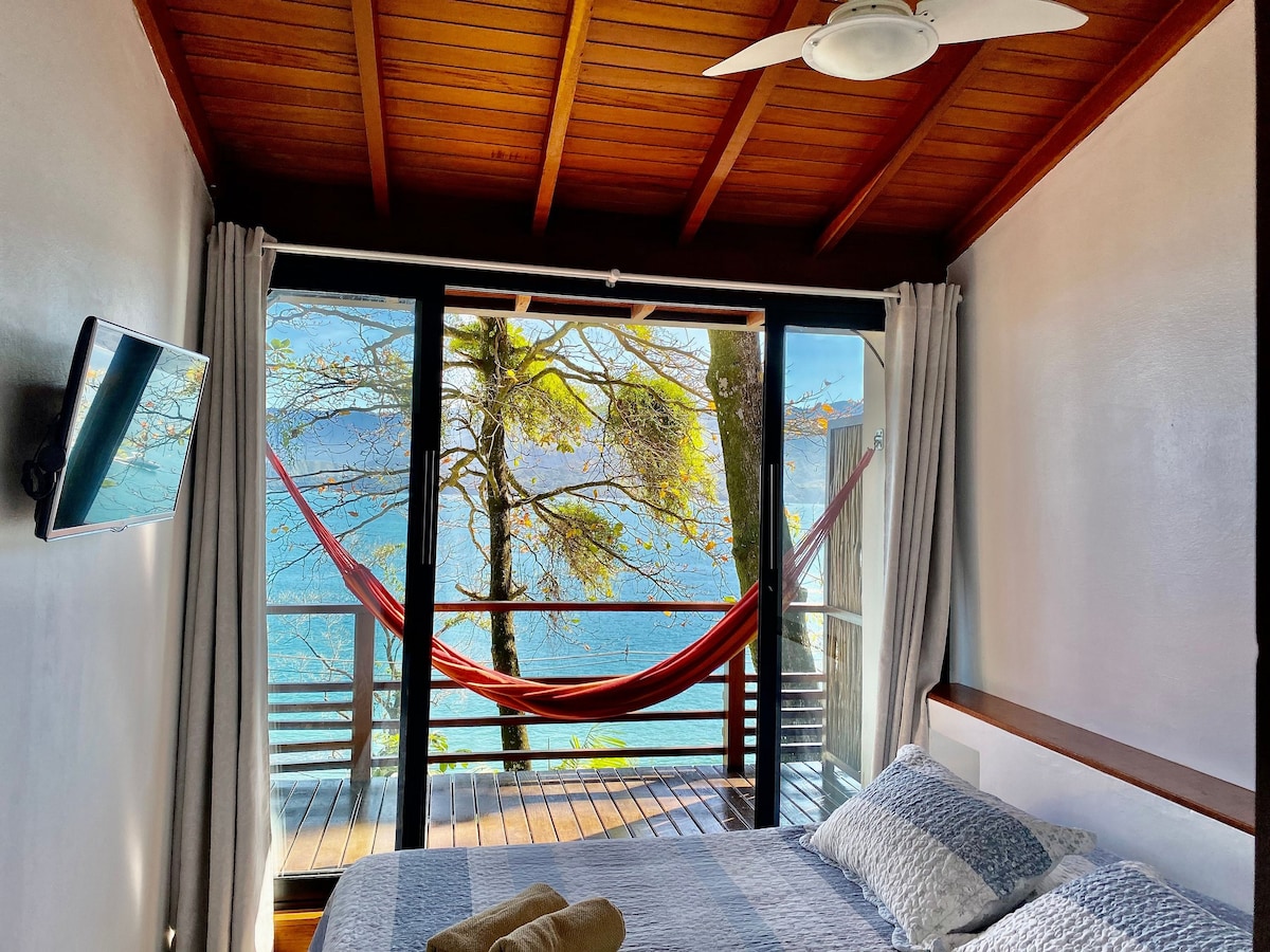 A well-lit bedroom features a spacious bed with neatly arranged linens. Large glass doors open to a private balcony adorned with a hammock, offering views of the surrounding trees and the tranquil sea beyond. A ceiling fan provides additional comfort.
