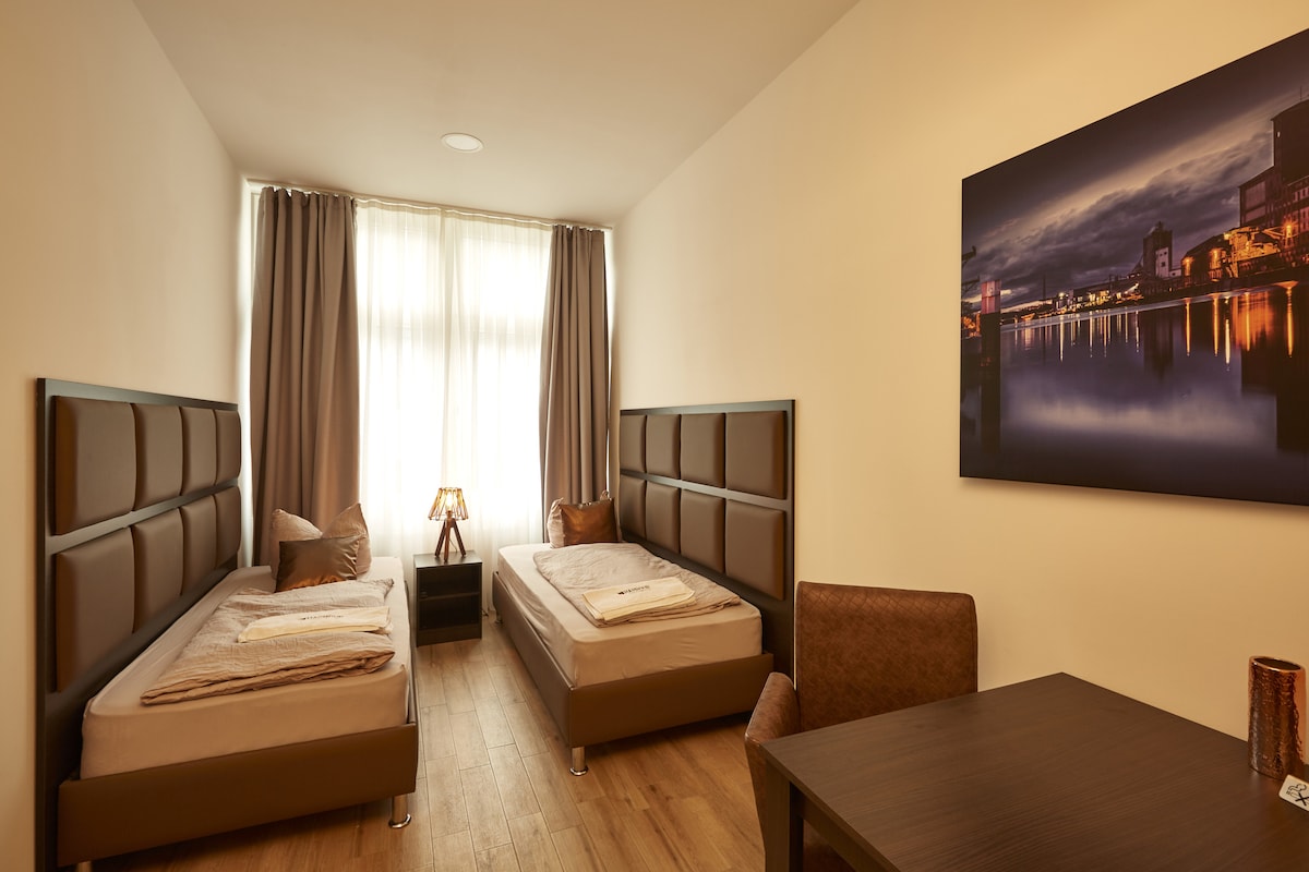 A light-filled room features two spacious beds separated by a small table. Decorative pillows are placed on each bed, while a desk and chair are positioned near the window. Floor-to-ceiling curtains frame the window, and a large artwork of a city skyline is displayed on the wall.