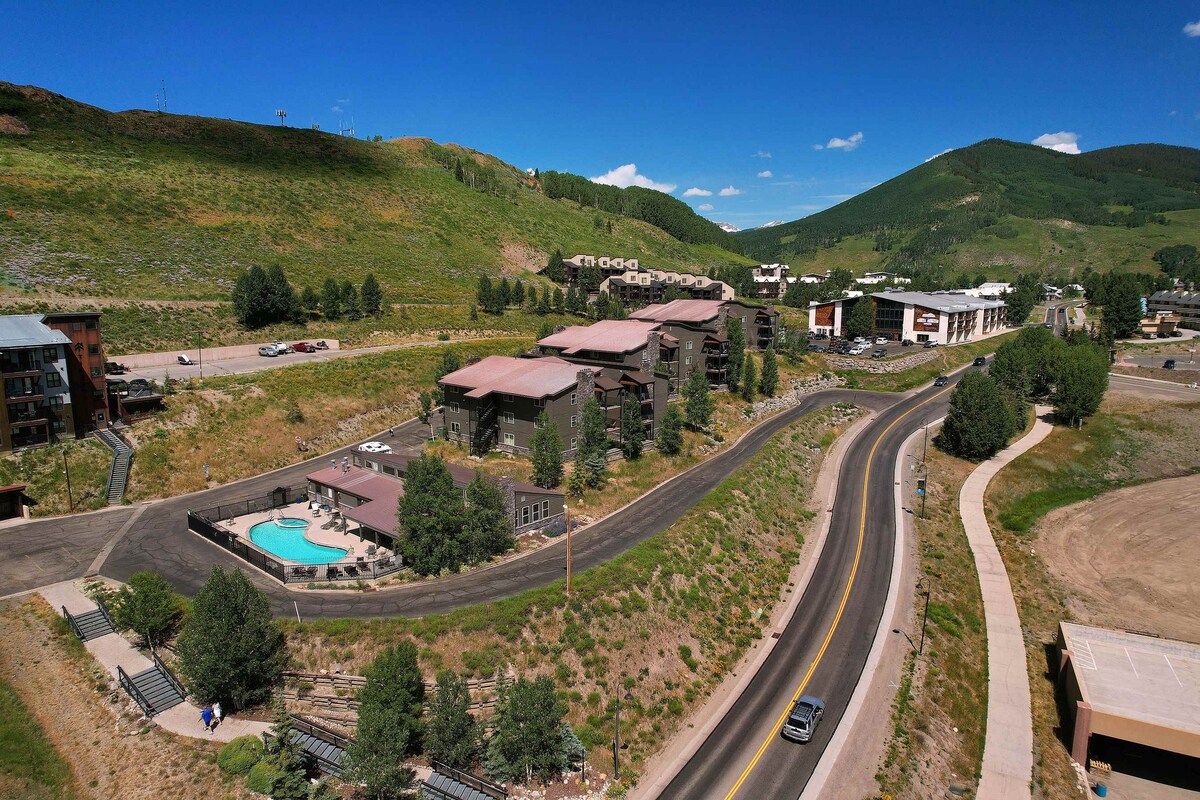 An aerial view of the Chateaux complex showcases its landscaped grounds and outdoor heated pool. The building's modern design is highlighted against a backdrop of lush green hills and blue skies, with the winding road leading to the property visible in the foreground.
