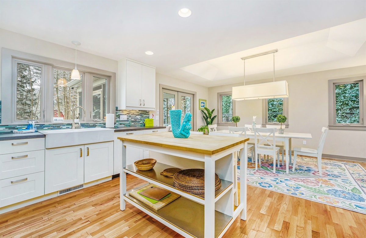 A bright kitchen features white cabinetry and stainless steel appliances, complemented by a central island with a wooden countertop. Natural light floods the space through multiple windows. A formal dining area is visible, showcasing a light-colored table surrounded by six chairs and decorative elements.