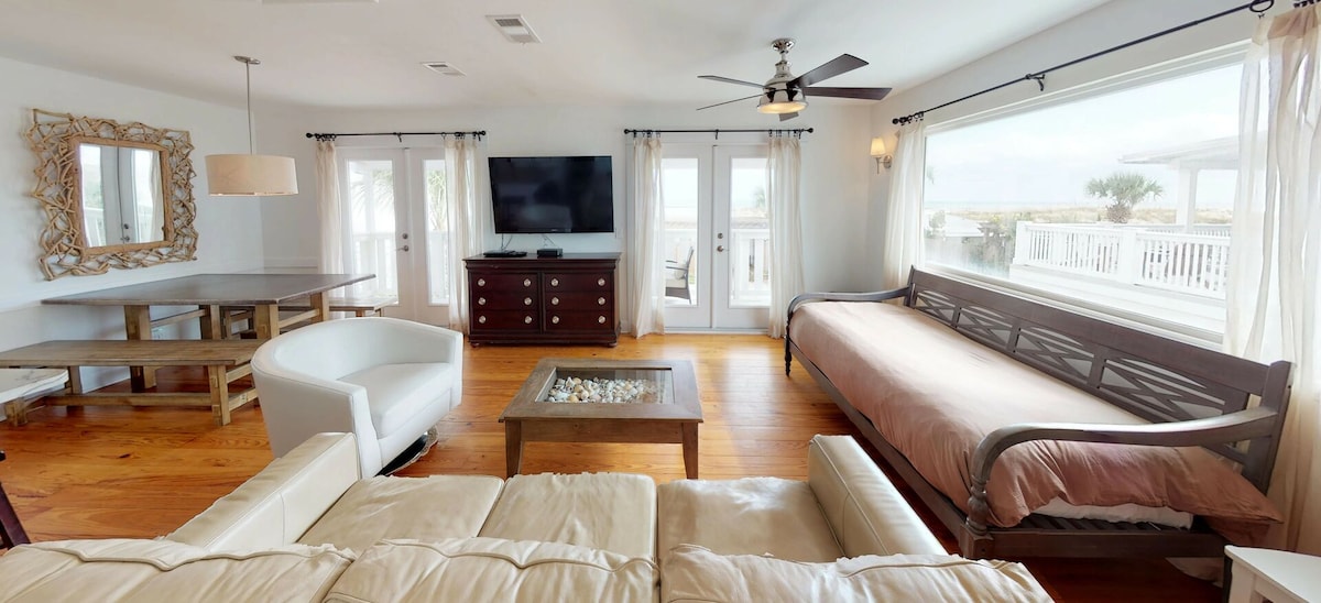 The Greatroom features Heart of Pine floors and abundant natural light from multiple windows. A large wall-mounted TV is visible, alongside a dining table and a cozy seating area with a sofa bed. French doors lead to an outdoor balcony, enhancing the inviting ambience.
