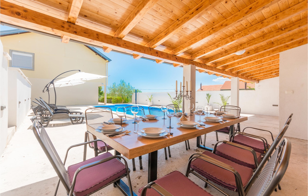 An outdoor dining area is set under a wooden pergola, featuring a large table surrounded by multiple chairs. The area is adjacent to a private pool, with sun loungers positioned nearby. Bright sunlight illuminates the space, creating a welcoming atmosphere for outdoor meals.