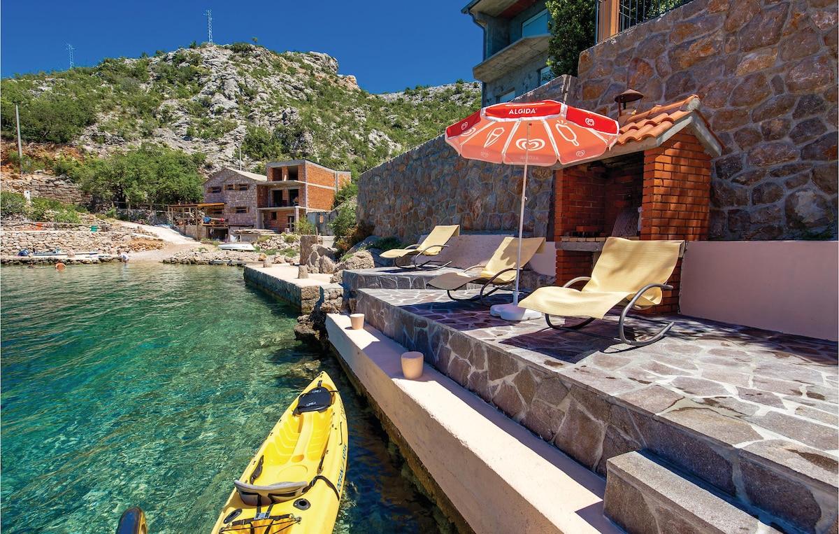 A concrete plateau beside clear water features two sun loungers under a red umbrella. A kayak is positioned nearby, while a stone wall adds texture to the tranquil setting. Lush greenery and a rocky hillside rise in the background, enhancing the natural landscape.