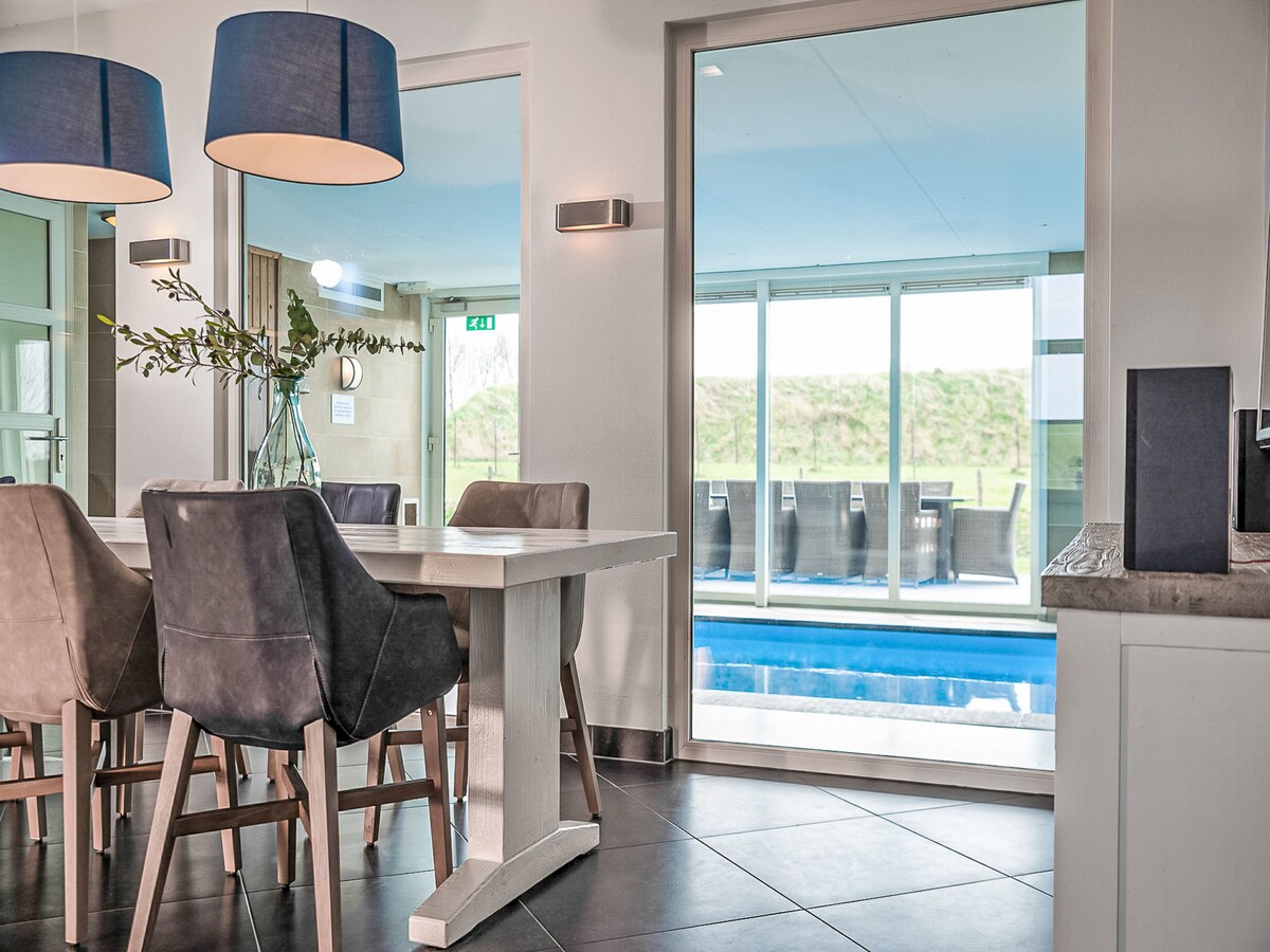 A modern dining area is presented, featuring a white table surrounded by dark upholstered chairs. Large glass doors provide a view of the swimming pool area, which is illuminated by natural light. The space is accentuated by two pendant lamps hanging above the table.