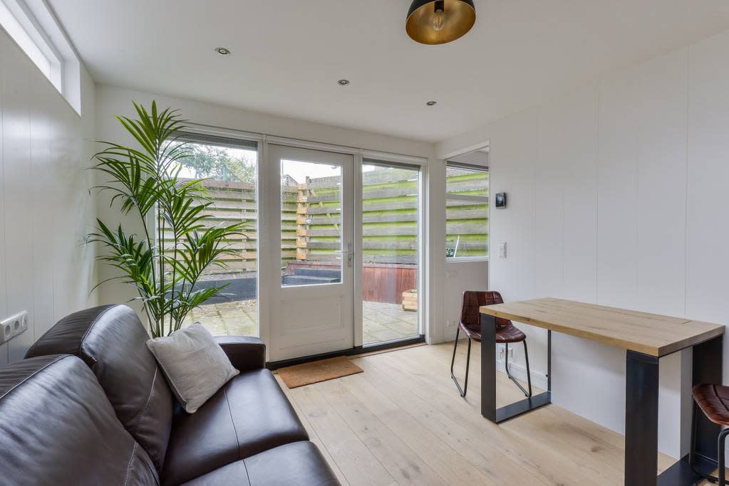 A modern living space features a dark leather sofa facing a simple wooden dining table with two stools. Large glass doors provide access to the private garden, while natural light brightens the interior. A tall plant adds a touch of greenery to the room.