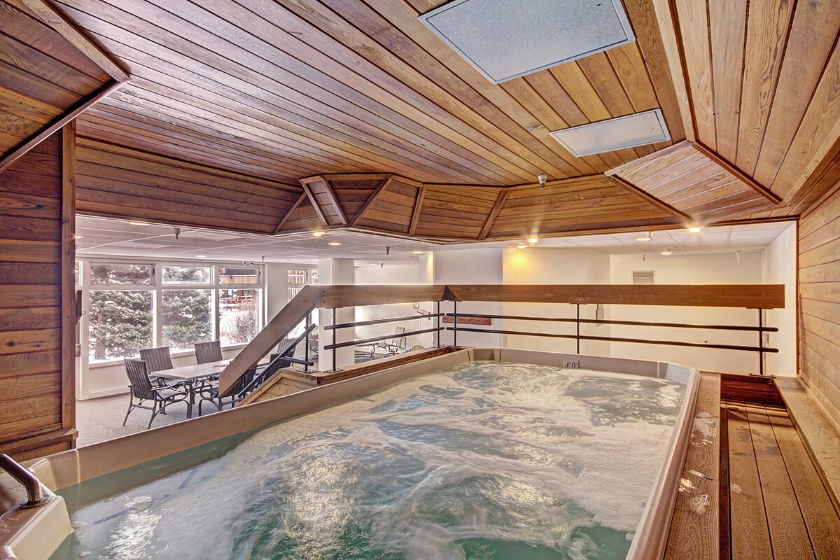 A shared indoor hot tub is situated in a well-lit space with wooden paneling and high ceilings. Large windows offer views of the outdoors. Nearby seating is provided by several chairs arranged for relaxation.