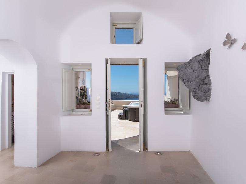 An entrance area is framed by two open windows, allowing natural light to brighten the space. A doorway leads outside, revealing views of the Aegean Sea and a private Jacuzzi area. The walls are minimally decorated, featuring a natural rock accent.