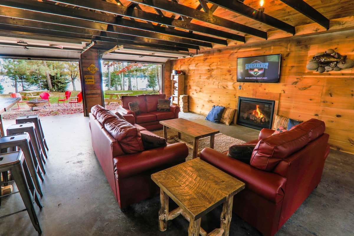 A spacious living area features comfortable leather sofas arranged around a wooden coffee table. A stone fireplace is located beneath a mounted television. Large garage doors provide a view of the outdoors, while barstools align against a wooden counter, enhancing the open layout.