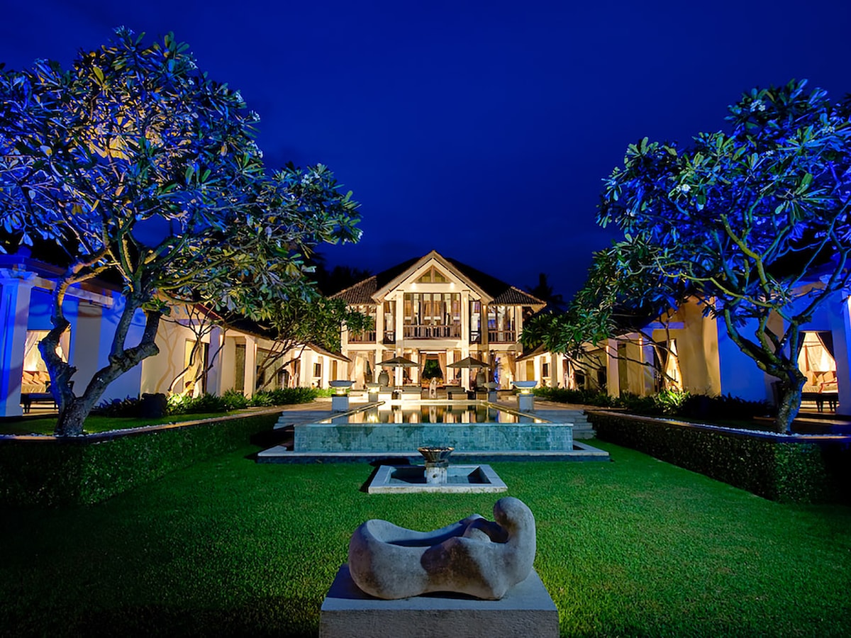 The villa's exterior is illuminated at night, highlighting lush greenery and artistic stone sculptures. A tranquil garden surrounds a central water feature, while the two-story pavilion showcases large windows reflecting the soft lighting, creating a serene and inviting ambiance.