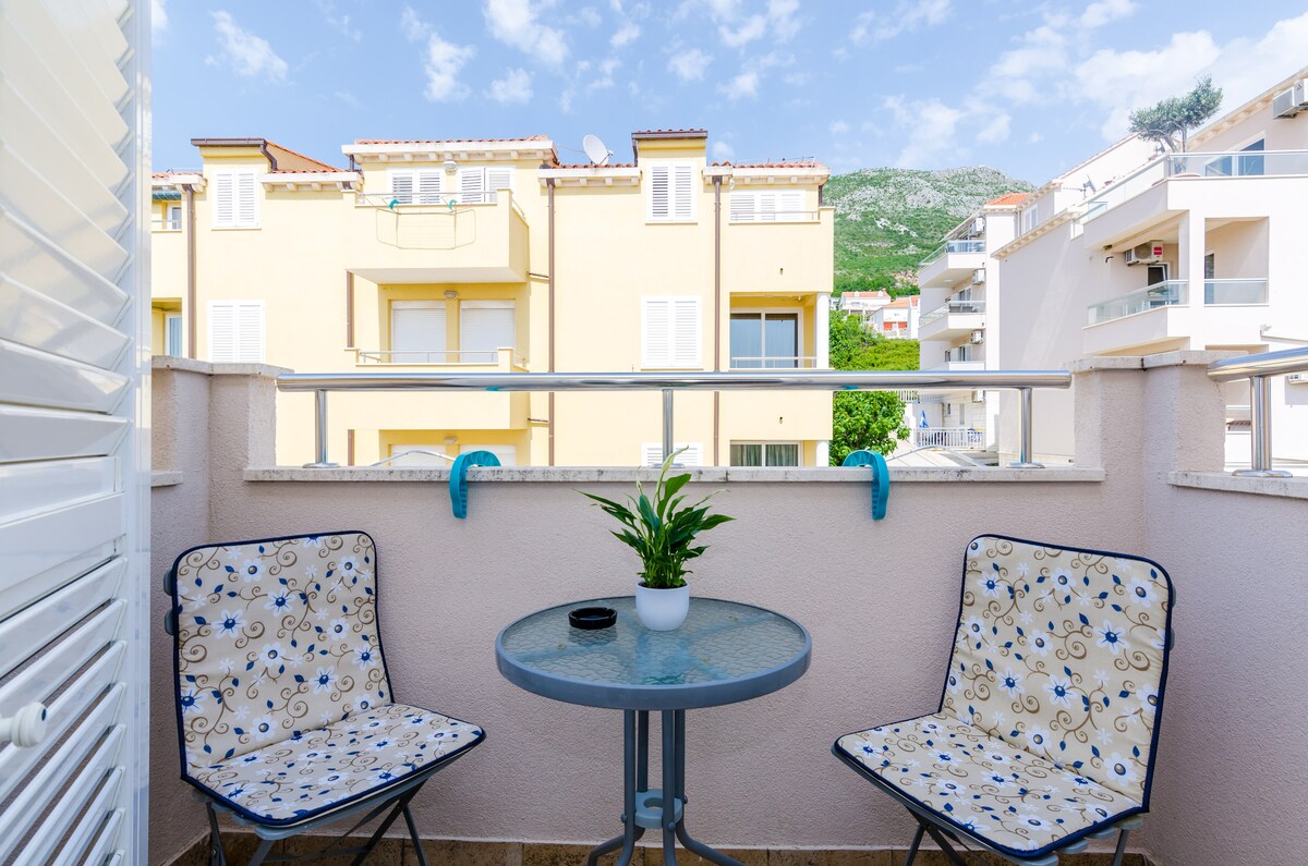 A furnished balcony is presented with two patterned folding chairs and a round table. A small potted plant is placed in the center of the table. The backdrop features neighboring buildings and a clear blue sky adorned with fluffy clouds.