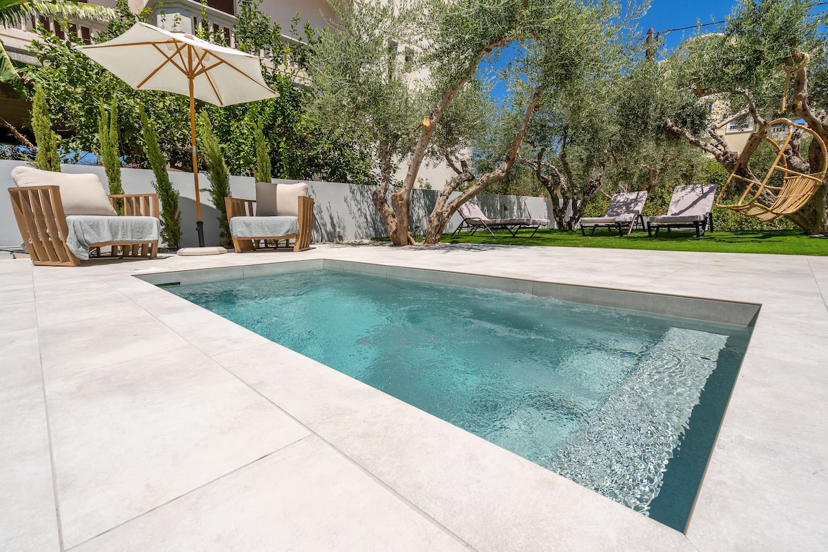 A private plunge pool is surrounded by a spacious deck featuring two wooden lounge chairs and a shaded umbrella. Olive trees create a serene backdrop, enhancing the tranquil outdoor setting.