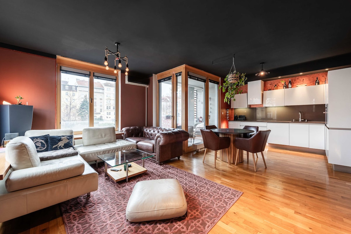 The living area features a blend of modern and cozy design, with a spacious leather sofa and a plush ottoman on a patterned area rug. A dining table with four chairs complements the fully equipped kitchen, while large windows bring in natural light, enhancing the ambiance.
