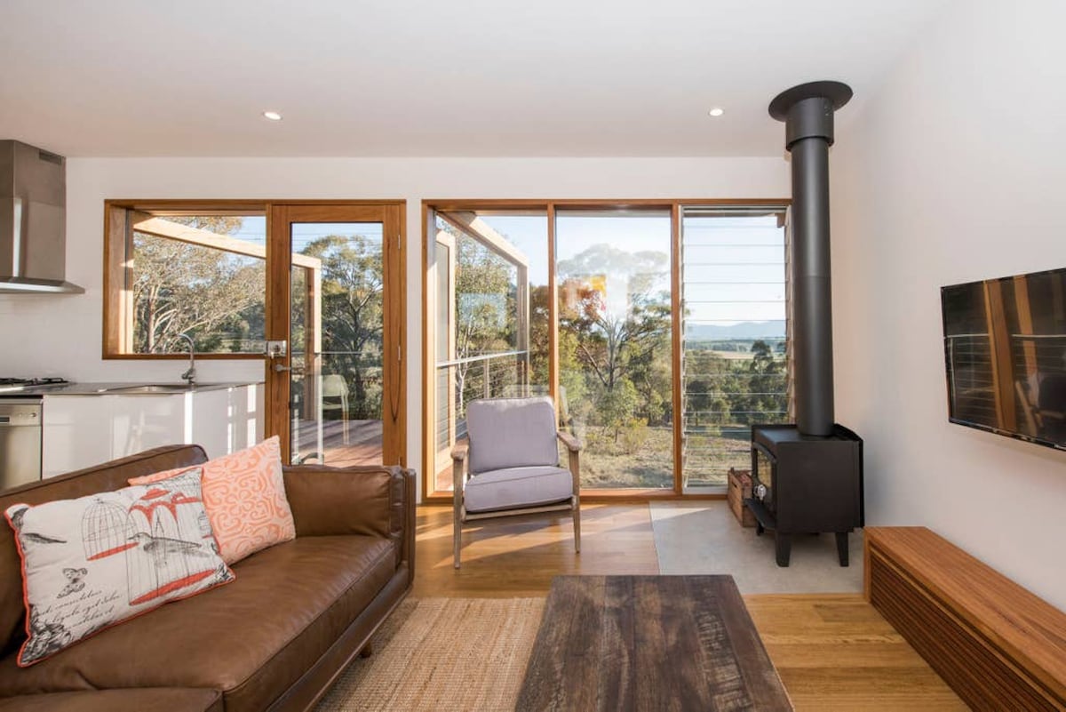 A cozy living area is highlighted by a comfortable brown sofa adorned with a decorative pillow. A modern chair is positioned nearby, with large glass doors revealing scenic views of the surrounding landscape. A wood stove and a flat-screen TV complete the space.