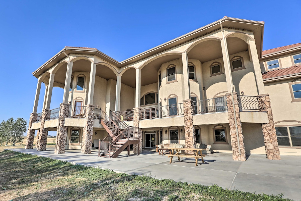 The exterior of a grand multi-level house features arched columns and stone accents. A spacious patio area is visible, complete with outdoor seating and a picnic table, while a staircase leads to the upper level. The surroundings are open and complemented by clear blue skies.