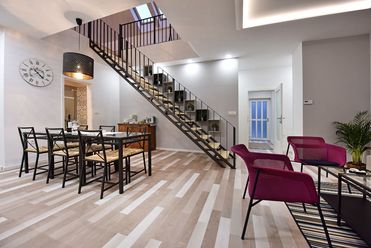 An open living space features a stylish dining area with a large table and six chairs. A sleek staircase leads to the upper gallery. Two modern armchairs are arranged near a coffee table, with natural light streaming through a skylight above.