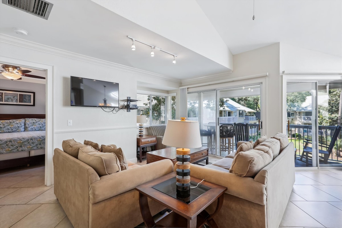 The living area is furnished with comfortable beige sofas arranged around a dark wooden coffee table. A wall-mounted 43" Smart TV is visible, and large glass doors allow natural light to fill the space while providing a view of the outdoor balcony and lush surroundings.