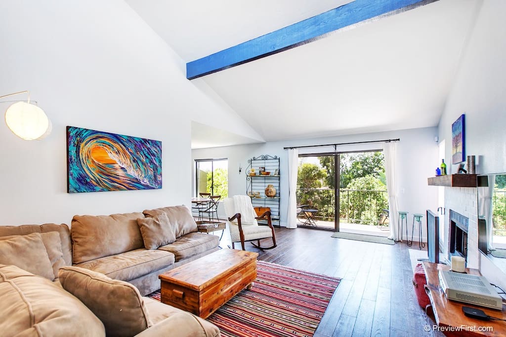 A spacious living area features a light-colored sectional couch and a wooden coffee table atop a colorful area rug. Large glass doors allow natural light to fill the room, leading to a view of greenery outside. A vibrant piece of wall art adds a touch of color.