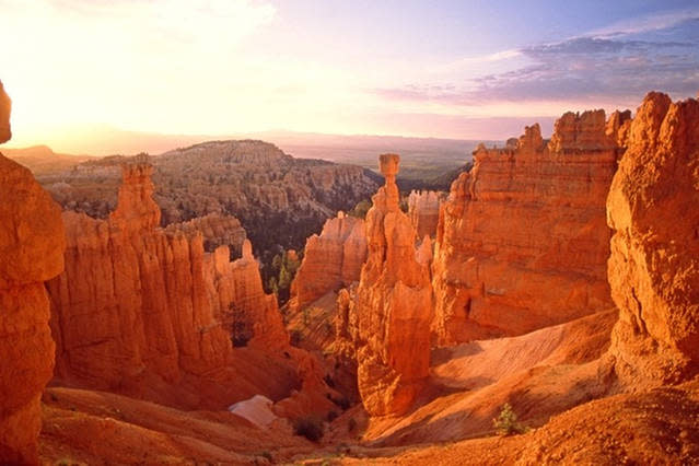 A panoramic view showcases the unique rock formations of Bryce Canyon, bathed in warm orange hues during sunset. The landscape features tall spires and hoodoos, surrounded by rugged terrain and distant hills under a softly lit sky.
