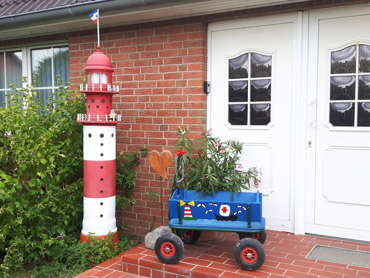 A brightly colored decorative lighthouse is positioned next to the entrance, featuring red and white stripes. Beside it, a blue wagon filled with vibrant flowers adds a cheerful touch to the brick facade, which is complemented by two white doors framed by bushes.