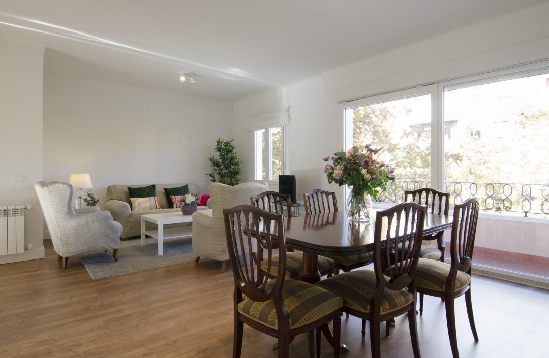 A welcoming living area features a cozy seating arrangement with two light-colored armchairs, a sofa, and a coffee table. A dining table with six chairs is illuminated by natural light from large windows, with floral arrangements enhancing the space's ambiance.