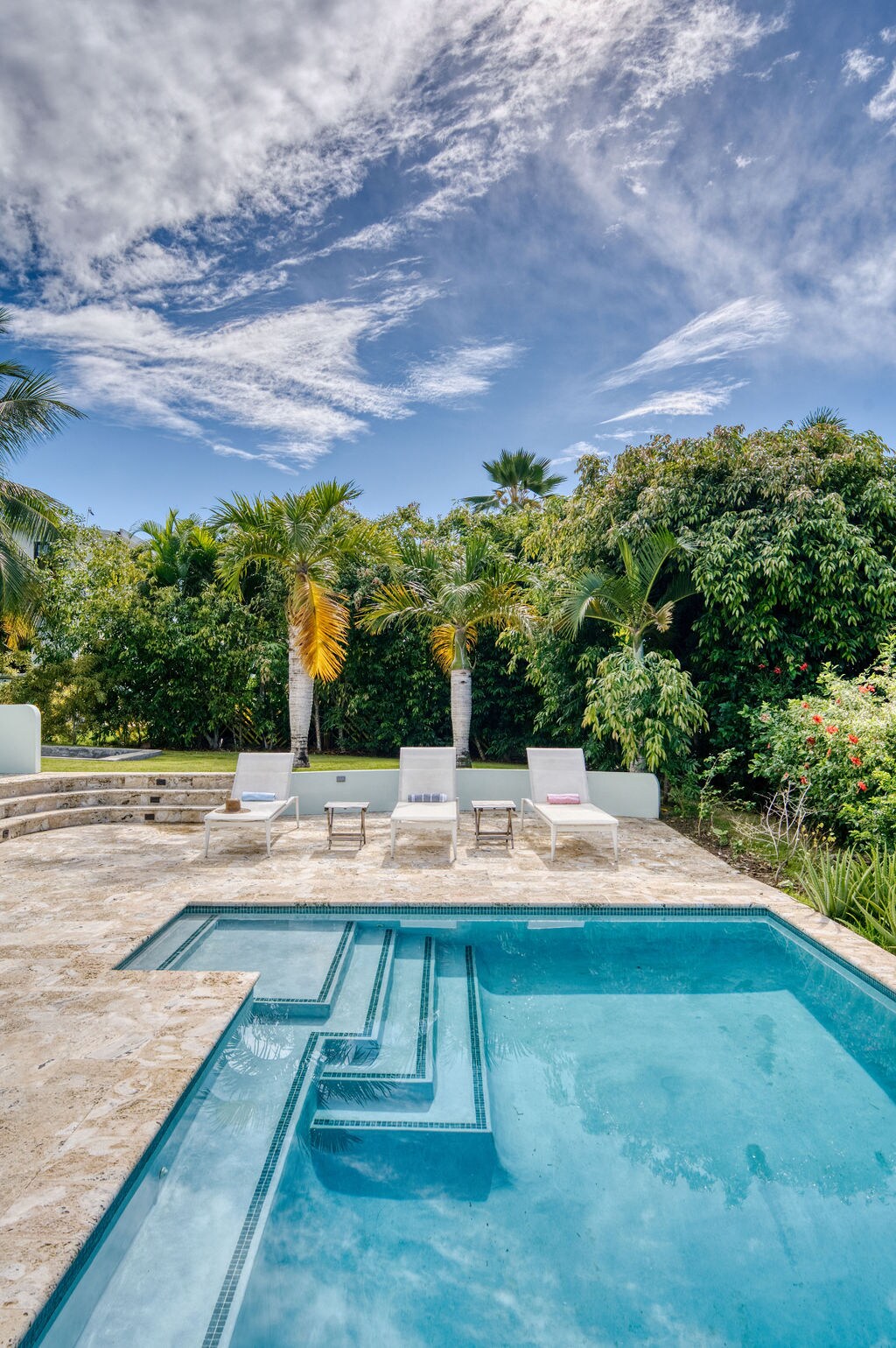 A private pool area is shown, featuring a clear blue pool with integrated steps. Surrounding greenery adds a natural backdrop, while several lounge chairs are positioned nearby. The scene is complemented by a bright sky interspersed with clouds, enhancing the outdoor ambiance.