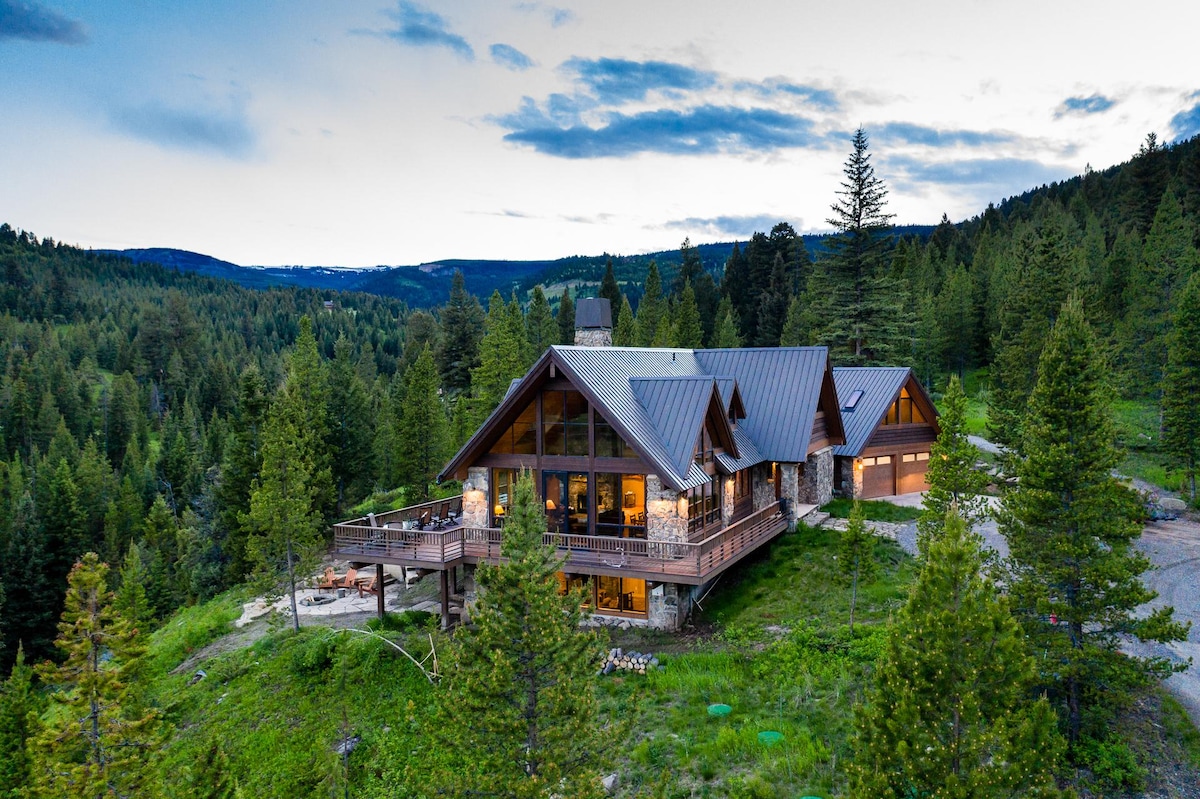 A spacious lodge is nestled among lush greenery, featuring large windows that provide a view of the wooded surroundings. The architectural style reflects a western design, with a blend of stone and timber finishes, and outdoor decking for enjoying the natural setting.