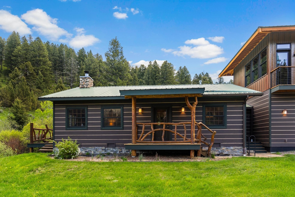 A charming cabin exterior is framed by lush greenery, highlighting a welcoming entrance with rustic wood railings. The cabin features a sloped metal roof and stone accents, surrounded by rolling hills and tall trees under a clear blue sky.