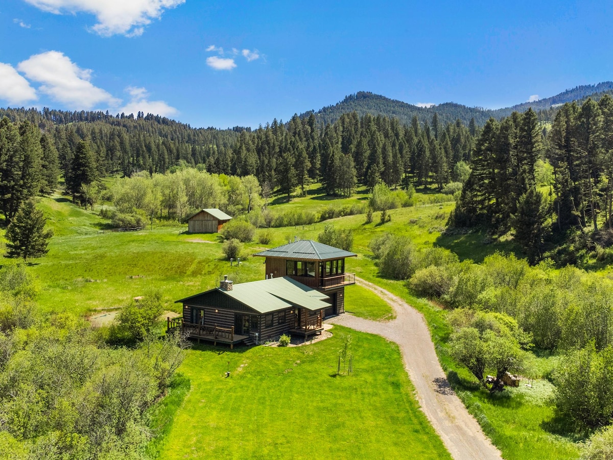 Aerial view of a rustic cabin situated on a lush green landscape, surrounded by trees and rolling hills. The cabin features a green metal roof and a wraparound porch, with walking paths leading to scenic nature. The tranquil setting showcases abundant foliage under a clear blue sky.