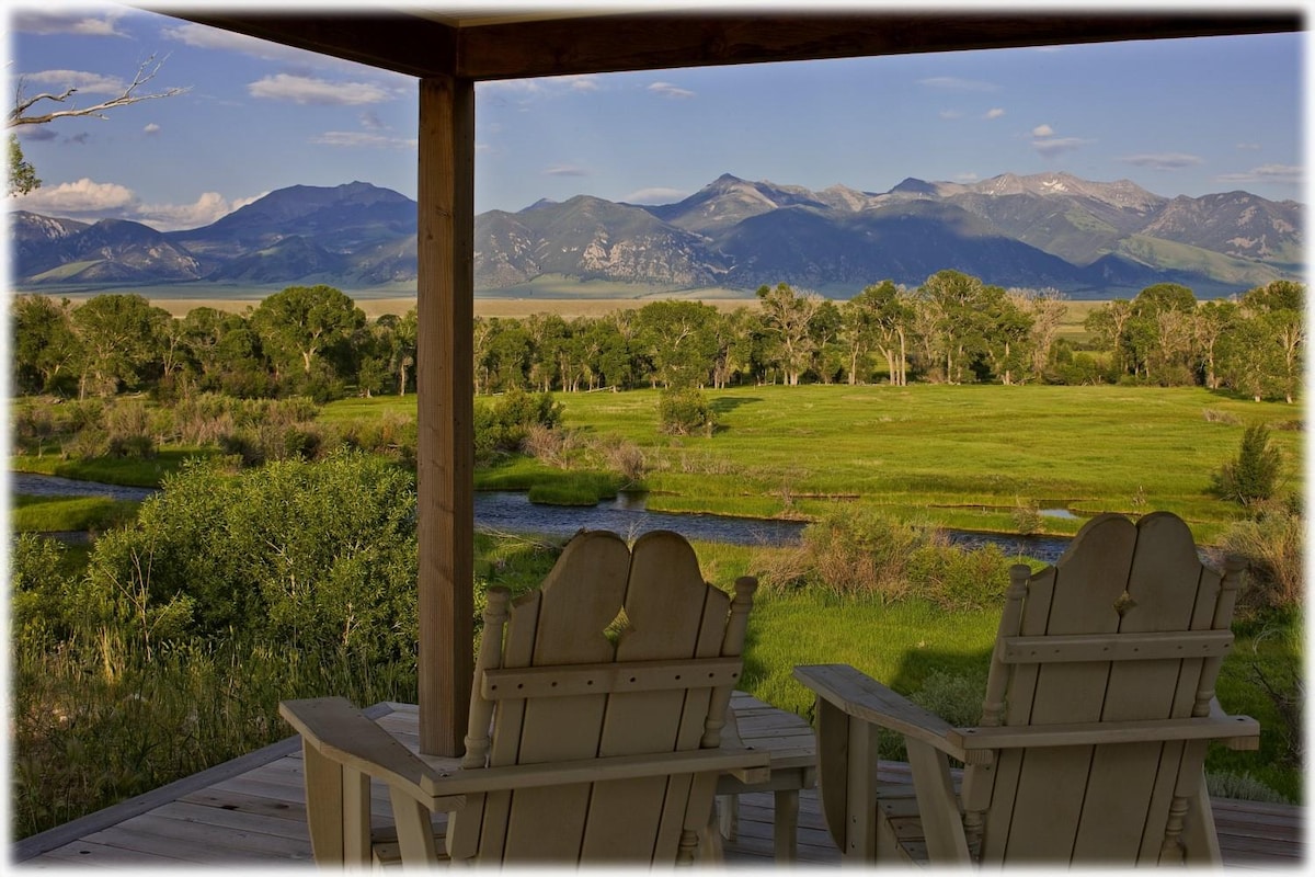 A pair of wooden adirondack chairs are positioned on a covered porch, overlooking expansive green fields that transition into a meandering river. The panoramic view includes distant mountains under a clear sky, enhancing the natural beauty of the Montana landscape.