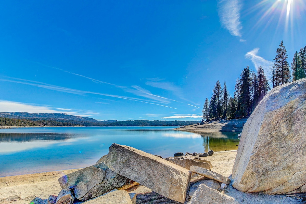 A serene lakeshore scene captures smooth rock formations leading to a calm body of water. Pine trees frame the background under a clear blue sky, with soft clouds gently streaking across. The tranquil atmosphere invites relaxation and connection with nature.