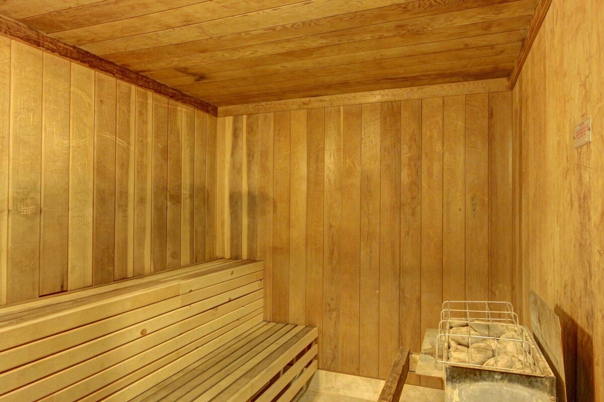 A warm wood-paneled sauna is presented, featuring slatted wooden benches positioned along the walls. The ceiling is also finished in wood, enhancing the natural ambiance. A small stone basin is visible in one corner, suggesting a space designed for relaxation.