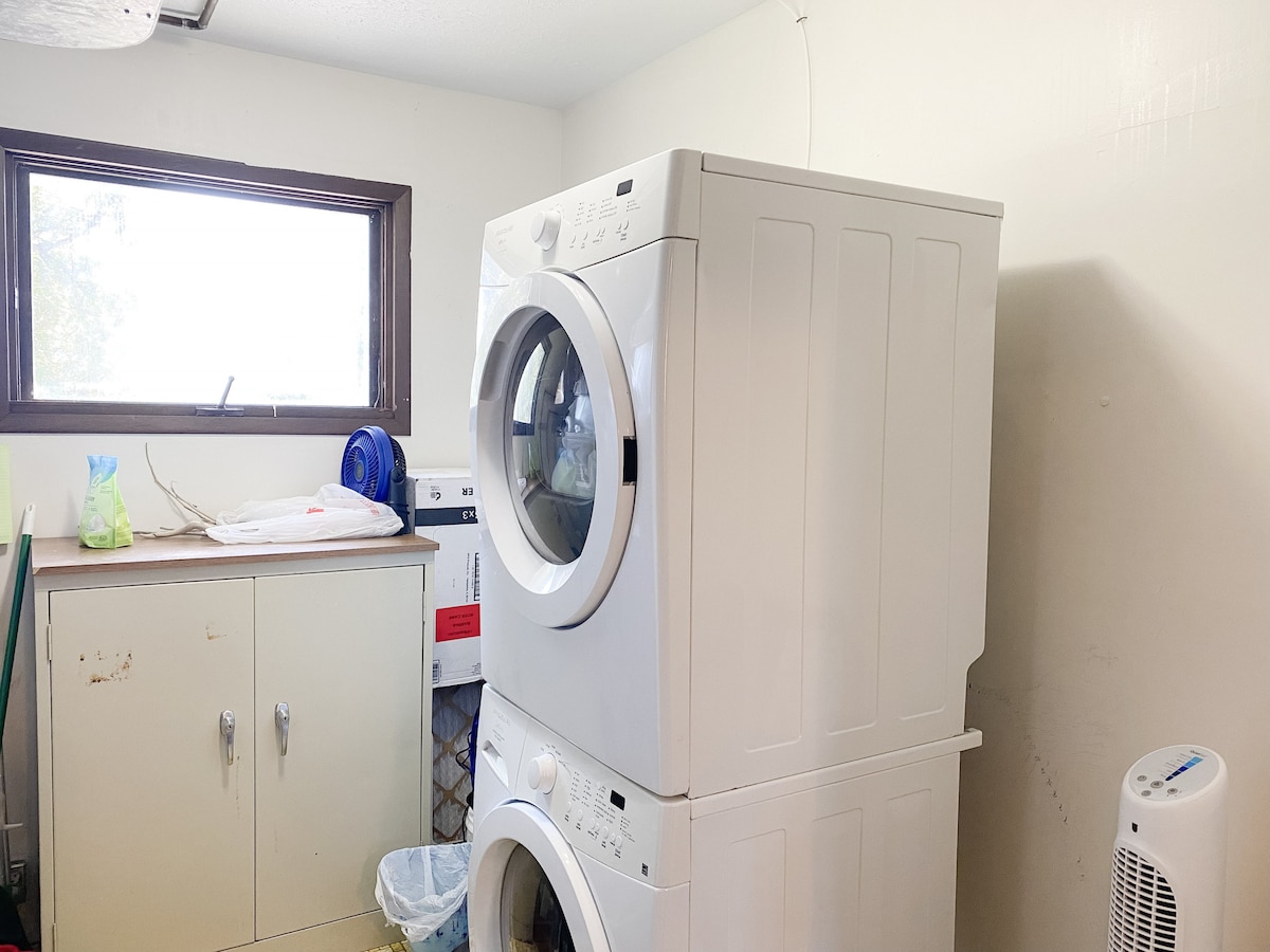 A laundry area is organized with a stacked washer and dryer, accompanied by a white cabinet. Natural light enters through a nearby window, illuminating the room. A wastebasket and additional cleaning supplies are visible, enhancing the functionality of the space.