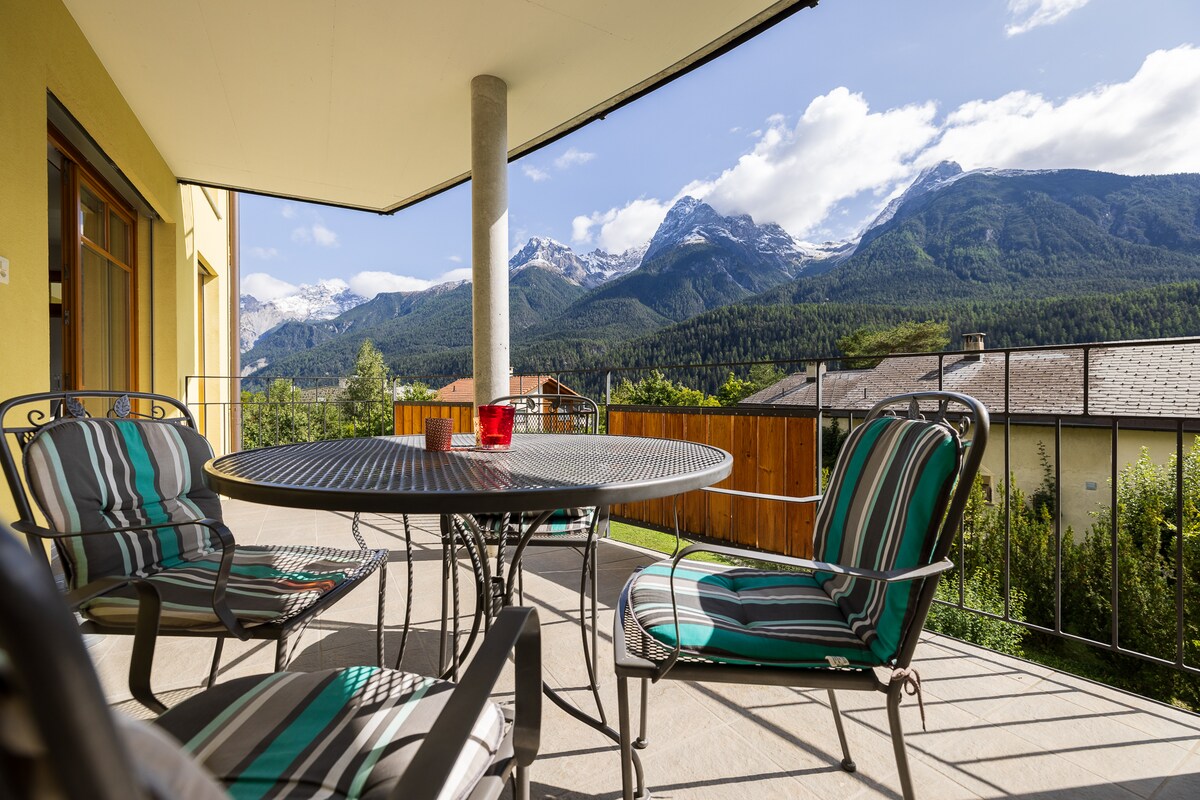 A spacious balcony features a round table surrounded by striped chairs, providing ample seating. The stunning panoramic views of mountains and greenery are visible in the background, while the bright sky enhances the tranquil outdoor space.